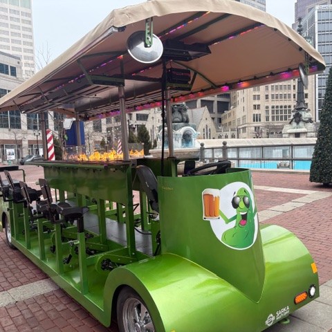 Green pedal pub vehicle with a cartoon pickle logo parked in an urban plaza.