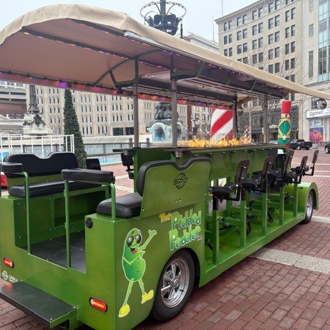 Green pedal-powered vehicle with cartoon pickle design in urban plaza setting.