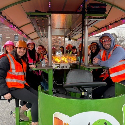 Group of people in orange vests and helmets on a pedal pub with a central fire feature.